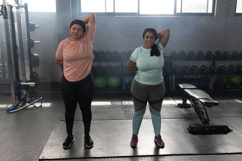 African american women stretching triceps together in gym weight room near dumbbells