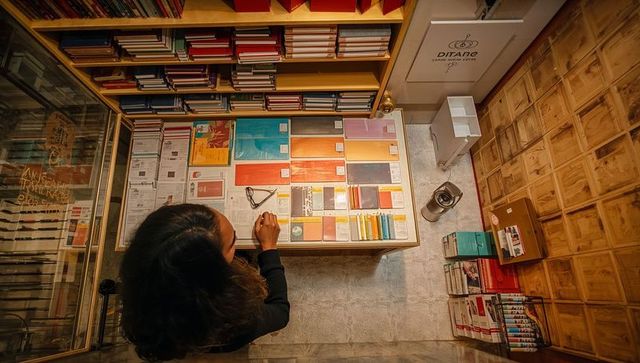 Overhead view of woman reaching colorful notebooks on wooden counter in stationery shop