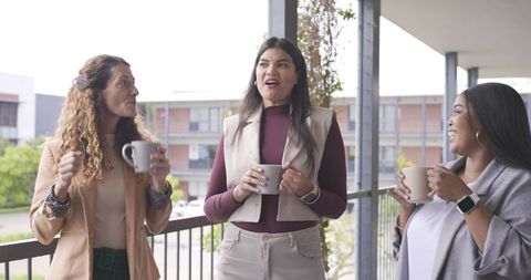 Diverse female coworkers chatting on balcony with coffee mugs, casual friendly outdoor break