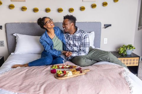 Romantic couple laughing and holding hands on bed with charcuterie and candlelight