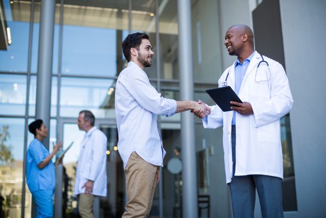 Doctor and patient shaking hands outside a modern hospital building. Medical professionals in the background engaging in conversation. Ideal for use in healthcare, medical services, patient care, trust in healthcare, and hospital promotional materials.