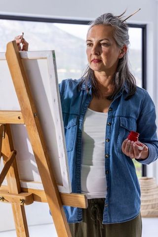 Senior woman painting on canvas in sunlit studio with lake view, creative art lifestyle