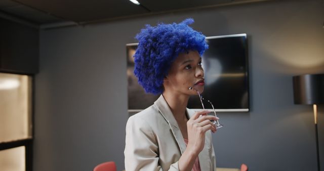 A woman with blue afro hair stands thoughtfully in a modern office while holding glasses. Ideal for themes related to workplace diversity, decision-making, creativity, business environments, and professional settings.