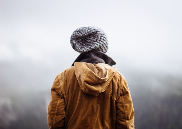 Person wearing a brown jacket and knit hat stands facing away in a foggy outdoor setting, possibly in a forest or mountainous area. Perfect for themes of solitude, contemplation, winter, and natural outdoor adventures.