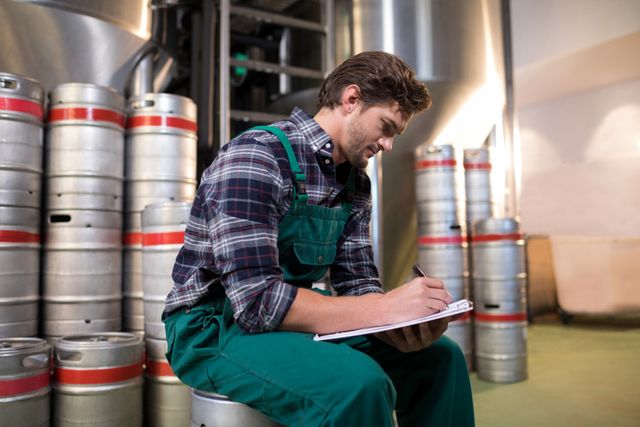 Warehouse worker in green overalls writing on paper while sitting on a keg in an industrial setting. Ideal for use in articles or advertisements related to logistics, inventory management, industrial work environments, and worker productivity.