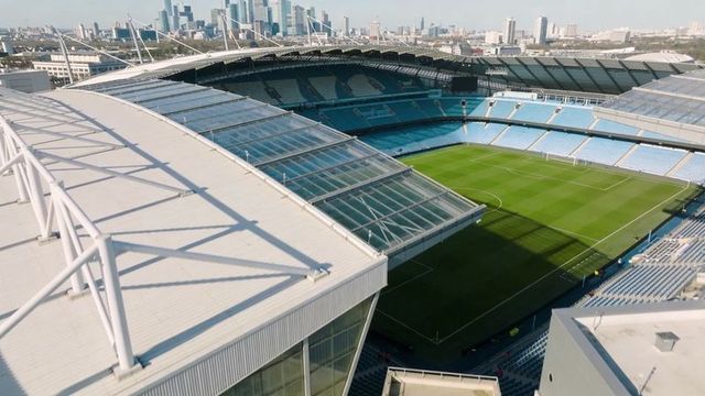 Aerial view of modern stadium with glazed canopy, striped pitch and light-blue seating