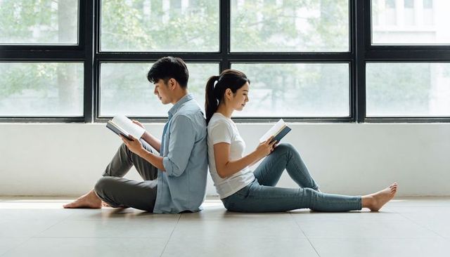 Young couple sitting back-to-back reading by large window in minimalist sunlit room