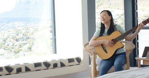Asian woman singing and playing acoustic guitar by bright window with mountain view