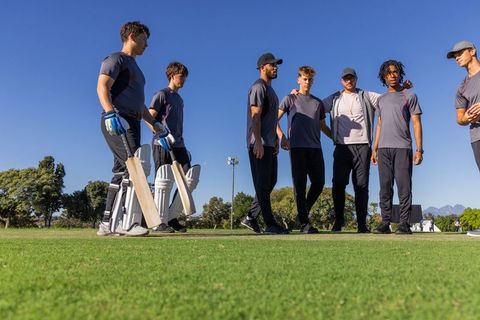 Diverse male cricket team standing and holding bats on sunlit pitch before match
