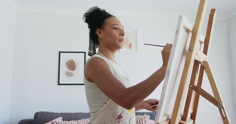Woman Painting on Canvas at Easel in Bright Home Living Room, Wearing Paint Apron