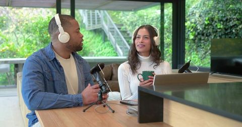 Diverse co-hosts recording home podcast with headphones, microphones and laptop in modern kitchen