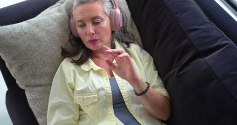 Mature woman relaxing on dark sofa listening to music with pink headphones