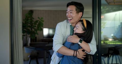 Asian couple laughing and hugging in modern home by sliding glass doors, joyful embrace