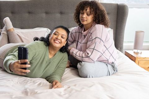 Diverse friends posing selfie on cozy bed wearing knit sweaters and loungewear in daylight