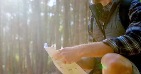 Crouching hiker consulting map in sunlit conifer forest with backpack and lens flare