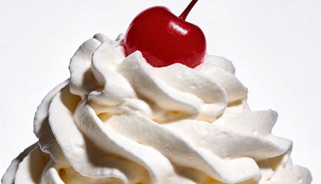 Whipped cream swirl topping with glossy red cherry close-up on white backdrop