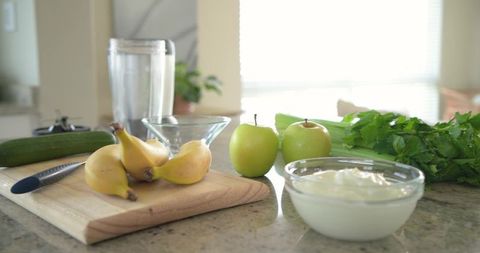 Sunlight streaming over countertop with bananas, green apples, yogurt and blender