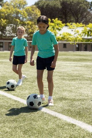 Girls practicing soccer dribbling on sunny school field wearing teal shirts and navy shorts