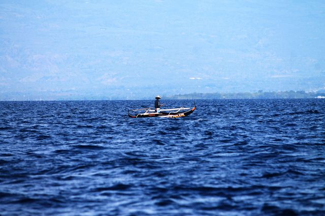 Image shows a lone fisherman in a small traditional fishing boat on a vast, clear blue ocean. Ideal for illustrating themes of solitude, traditional fishing methods, maritime activities, tranquility, travel, and remote living. Suitable for use in articles, travel brochures, maritime industry ads, and educational materials about sustainable fishing practices.