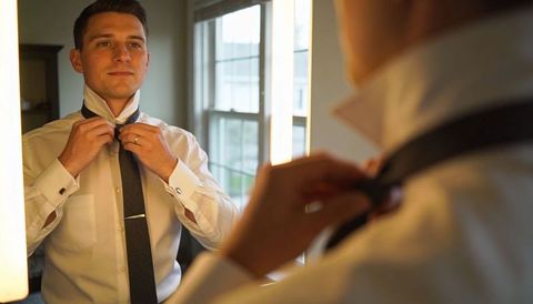 Man adjusting tie in mirror with warm vanity lights preparing formal outfit
