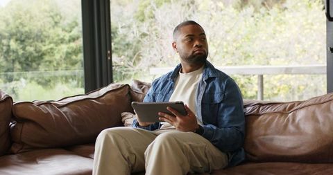 African american man sitting on leather couch holding tablet looking out window