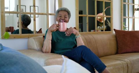Senior African American woman relaxing on leather sofa holding pink mug, smiling at home