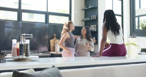 Three women chatting around sunlit modern kitchen island in casual athleisure