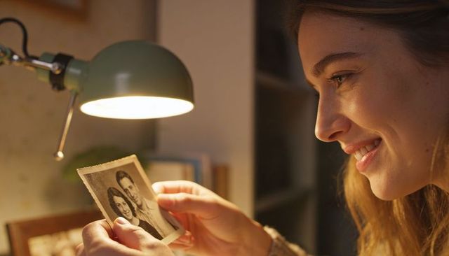 Young woman holding vintage black-and-white portrait under warm desk lamp smiling with nostalgia