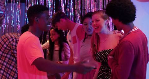 Young friends laughing and chatting under magenta-blue party lights with glitter fringe