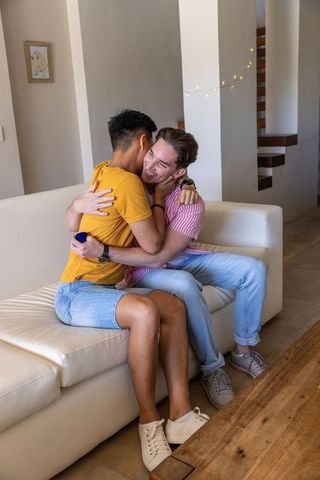 Diverse male couple embracing on cream sofa holding blue ring box celebrating proposal