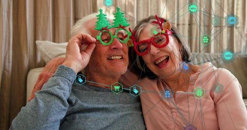 Senior couple wearing christmas glasses laughing on sofa with e-commerce network overlay