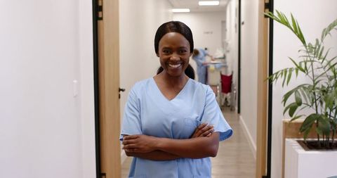 Smiling nurse standing with arms crossed in clinic corridor wearing light blue scrubs