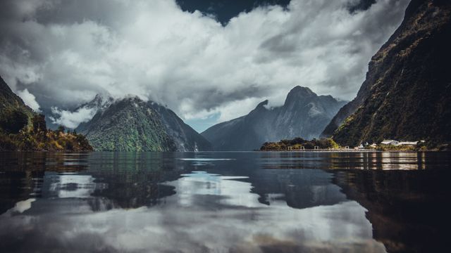 This stock photo captures a serene, mountainous landscape with clouds reflecting on calm lake waters, creating a mirror-like effect. Ideal for marketing materials related to travel, nature, outdoor adventures, and environmental conservation. Perfect for use in websites, posters, brochures, and social media campaigns promoting scenic destinations and eco-tourism.