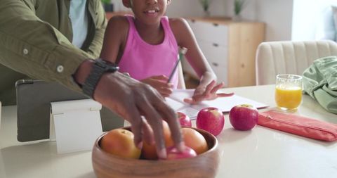 Parent helping child studying at sunlit kitchen table, fruit bowl, apples and juice