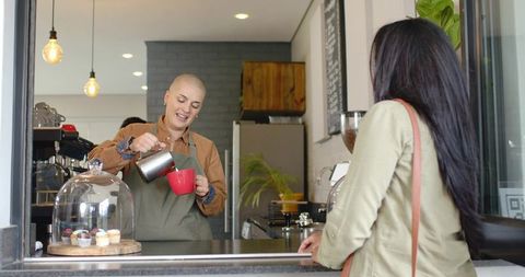 Barista pouring milk into red cup at cafe window with indian customer waiting and smiling