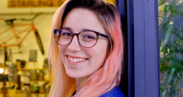 Young woman with pink hair and glasses smiling while indoors. This image can be used for promotional materials, lifestyle blogs, fashion articles, or content related to youth culture and joy.