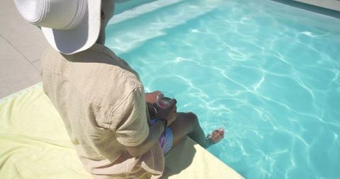 African american man relaxing poolside with wide-brim hat and cool drink, dipping toes