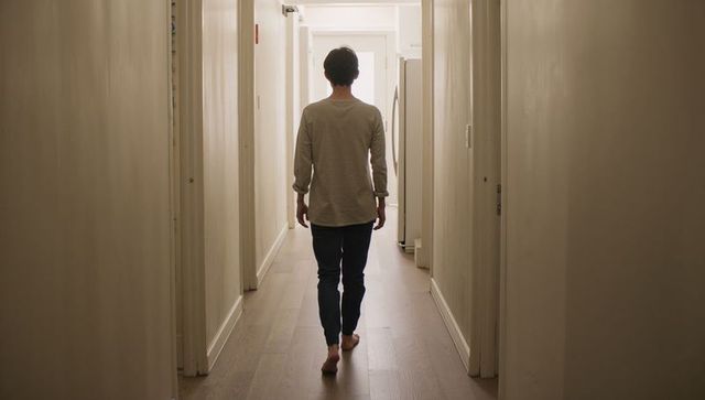 Barefoot man walking down narrow backlit hallway toward bright doorway in home interior