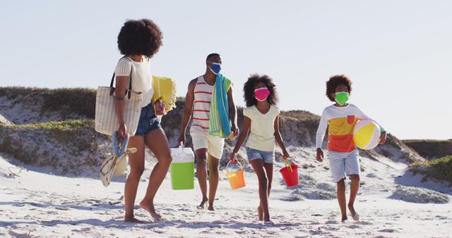 Image shows a cheerful family of four, including parents and two children, walking on the beach while carrying sand toys and beach equipment, with everyone wearing protective masks. Perfect for topics on family vacations, beach trips, safe travel, summer activities, and outdoor fun. Ideal for travel websites, family and lifestyle blogs, health and safety promotions, and tourism brochures.
