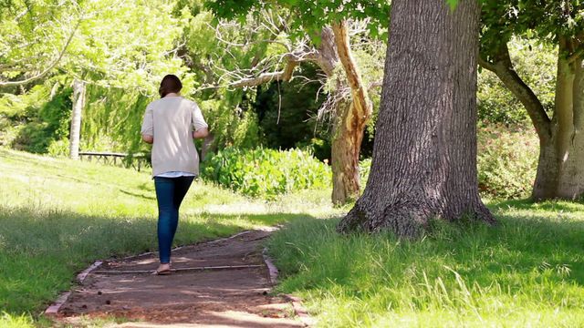 Young woman walking alone in a park on a sunny day. The environment is lush and green, with tall trees and a well-worn path. Ideal for concepts like relaxation, natural beauty, mental health, and personal time. Suitable for use in articles or campaigns related to wellness, nature conservation, and outdoor activities.