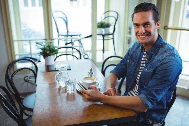 Young smiling man sitting at a wooden table in a cozy cafe, using his mobile phone. He is wearing a casual denim jacket and striped shirt. The cafe setting includes a cup of coffee, water glass, and natural light coming through large windows, creating a relaxed atmosphere. Ideal for use in advertisements, social media, lifestyle blogs, or promoting cafes, casual dining, and technology products.
