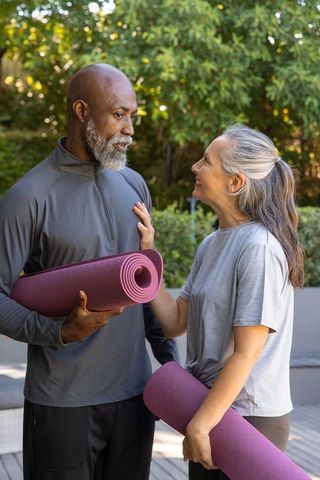 Diverse senior couple holding purple yoga mats and smiling before outdoor yoga session