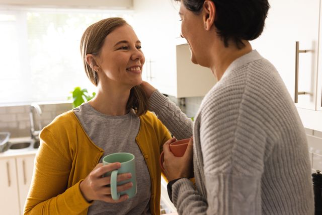 This image shows a mid adult lesbian couple smiling and looking at each other while holding coffee mugs in a kitchen. It can be used for themes related to love, romance, togetherness, and domestic life. Ideal for articles, blogs, or advertisements focusing on LGBTQ+ relationships, home life, and morning routines.