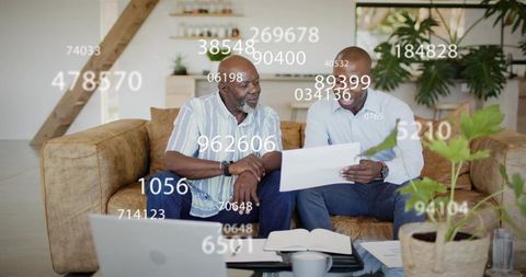 Senior and adult men reviewing financial documents on sofa with laptop and data overlay