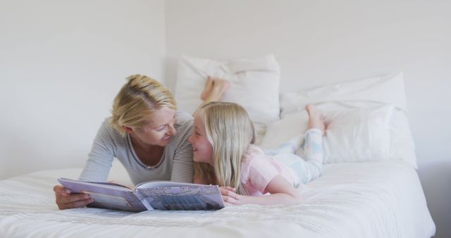 Happy caucasian mother and daughter ready story book together lying on bed at home. Motherhood, childhood, fun, togetherness and domestic life.