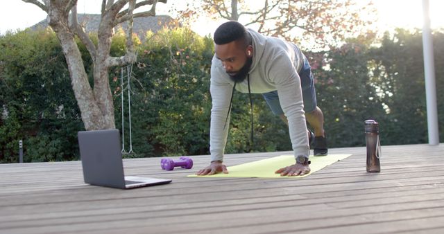 Man wearing workout attire performing plank exercise outdoors on a yellow yoga mat, following an online session on a laptop. Purple dumbbell and water bottle beside him. Ideal for illustrating topics about home workouts, fitness routines, outdoor exercise, staying active during quarantine, and online fitness coaching.
