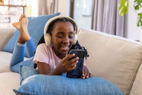 Smiling african american girl lying on couch wearing headphones and using smartphone