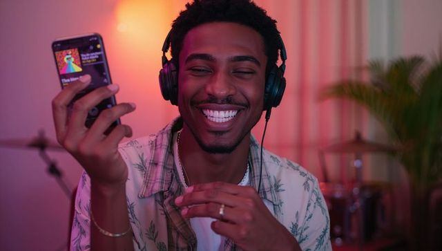 Smiling man with headphones holding smartphone showing album art in cozy music studio