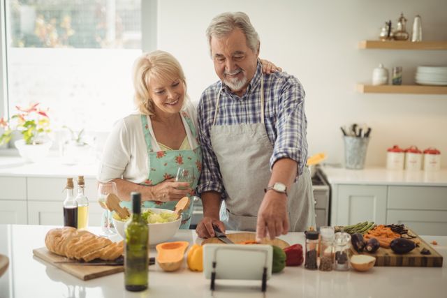 Senior couple preparing a healthy meal in a modern kitchen. They are smiling and enjoying each other's company while chopping vegetables and making a salad. The scene includes fresh ingredients like bread, wine, and various vegetables. Ideal for use in advertisements, articles, or blogs about healthy living, senior lifestyle, family bonding, and home cooking.