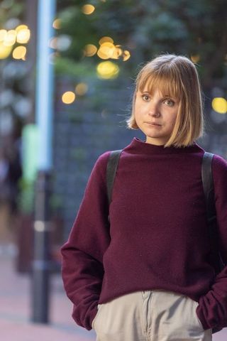Young woman standing on city street at dusk wearing burgundy sweater and backpack